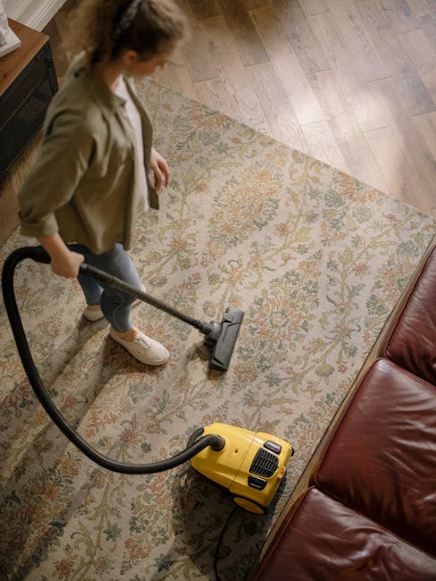 A woman using a yellow vacuum cleaner to deep clean a patterned area rug with floral motifs in a living room, featuring wooden flooring and a brown leather sofa. The room is well-lit, and the surface 