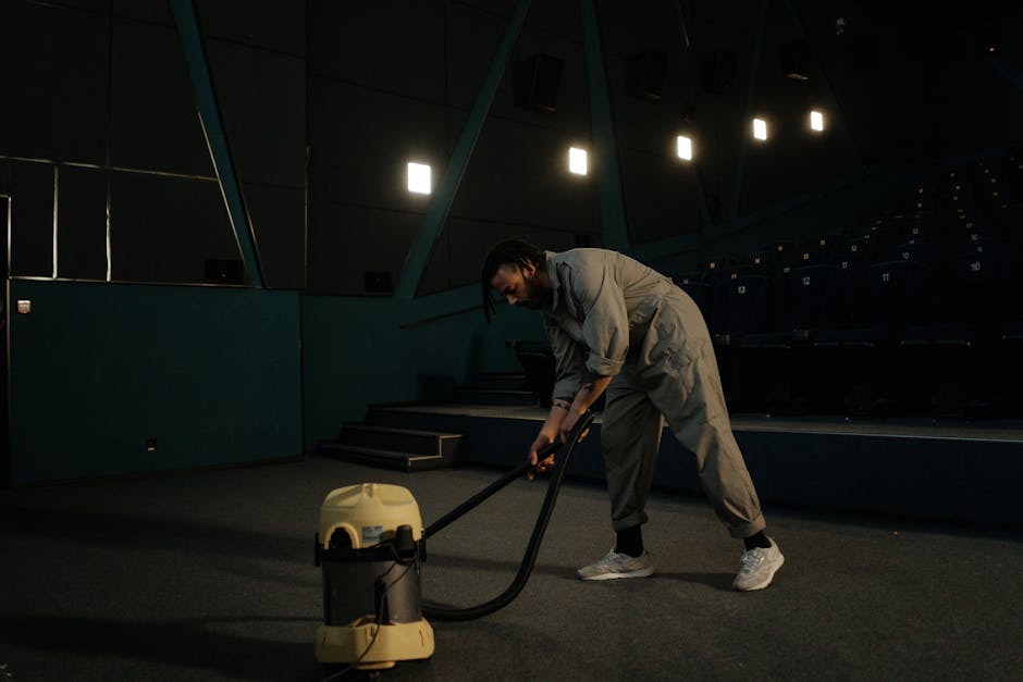 A person cleaning a beige textured area rug in a living space with a red upright vacuum cleaner labeled with words related to upholstery and deep cleaning, positioned on the rug. The individual is bar